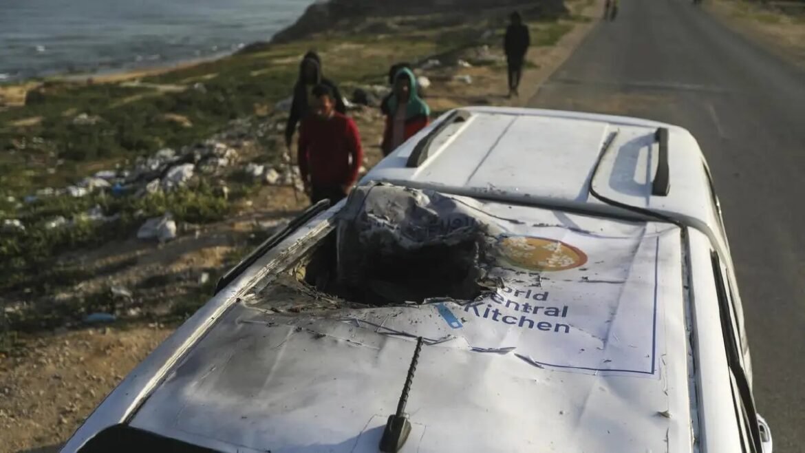 White van with a large hole in the roof, displaying a World Central Kitchen logo, on a coastal road with onlookers nearby.