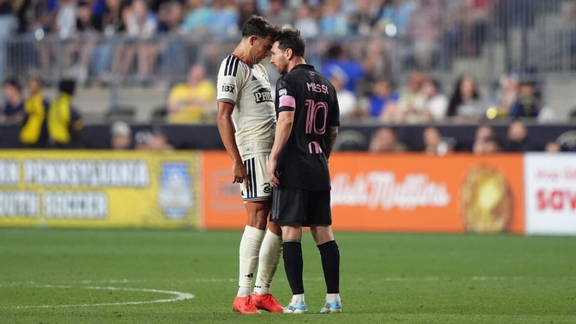 Two soccer players in contrasting kits stand nose-to-nose on the pitch during a match with a blurred crowd behind them.