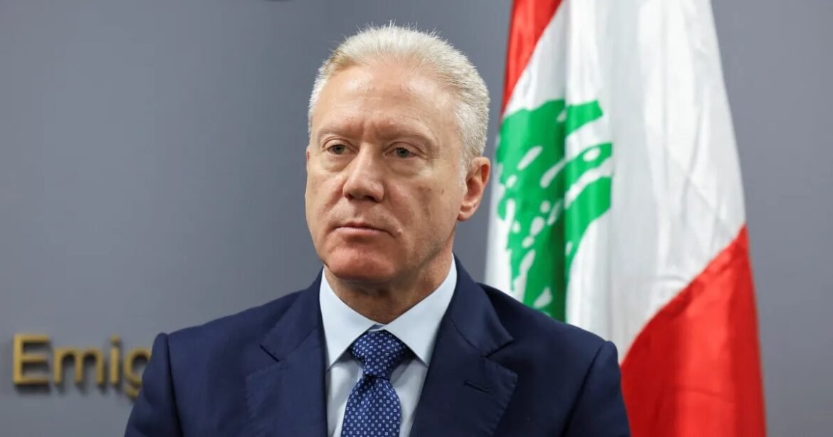 Older man in a navy suit and blue tie speaks at a formal event, with a red‑white‑green flag behind him.