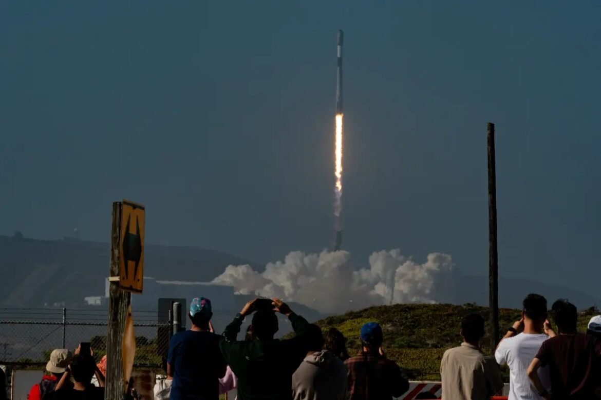 Crowd of spectators watching a rocket launch at dusk, the rocket ascending with a fiery plume above a smoky trail from the pad.