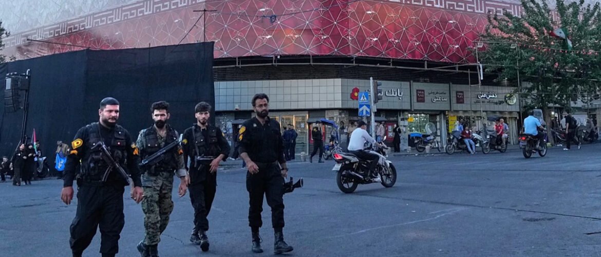 Group of four armed men in black tactical outfits walking across a city street with shops and motorbikes in the background.