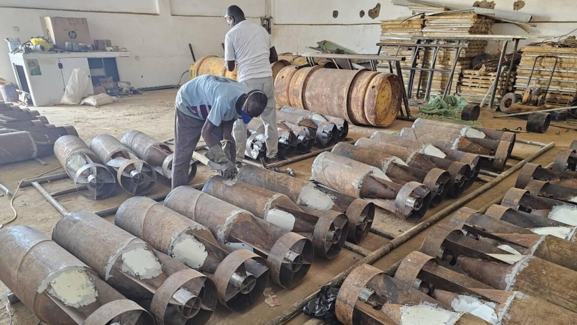 Two workers in a workshop inspecting or assembling a row of large rusted screw augers laid out on the floor, with tools and metal drums in the background.
