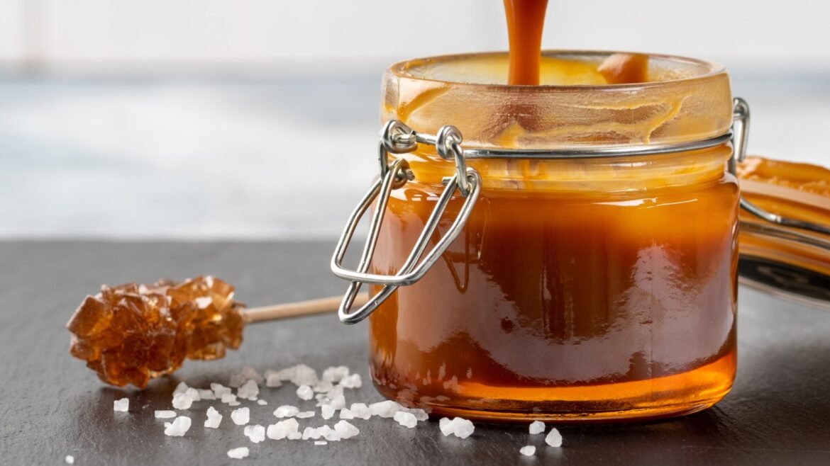 Glass jar of honey with a metal clasp, honey pouring from a wooden dipper beside scattered rock sugar on a dark surface.