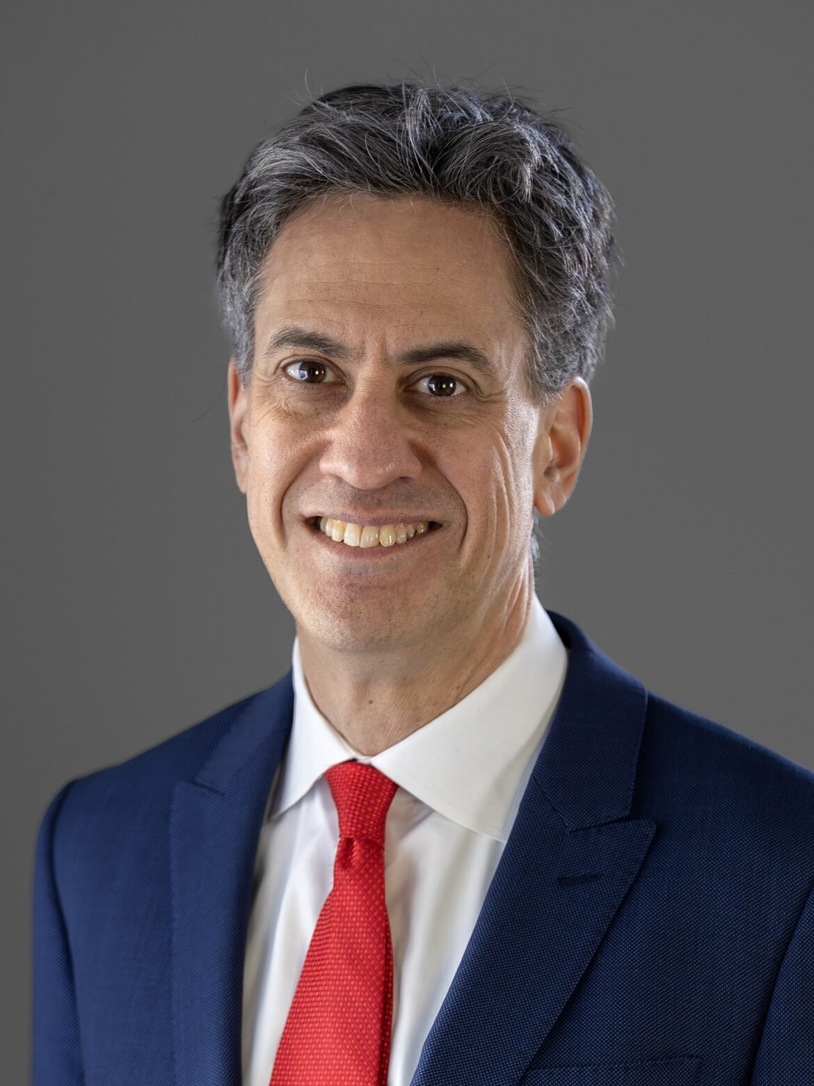 Smiling man in a blue suit and red tie against a gray background.