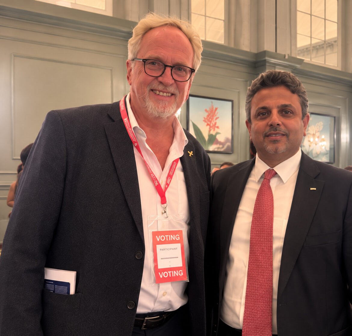 Two men in suits posing together at a voting event, one wearing a red lanyard and badge that reads VOTING.