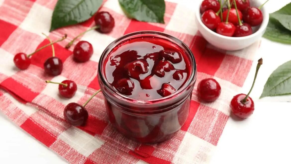 Glass jar of cherry jam on a red-and-white checkered cloth with fresh cherries scattered around.