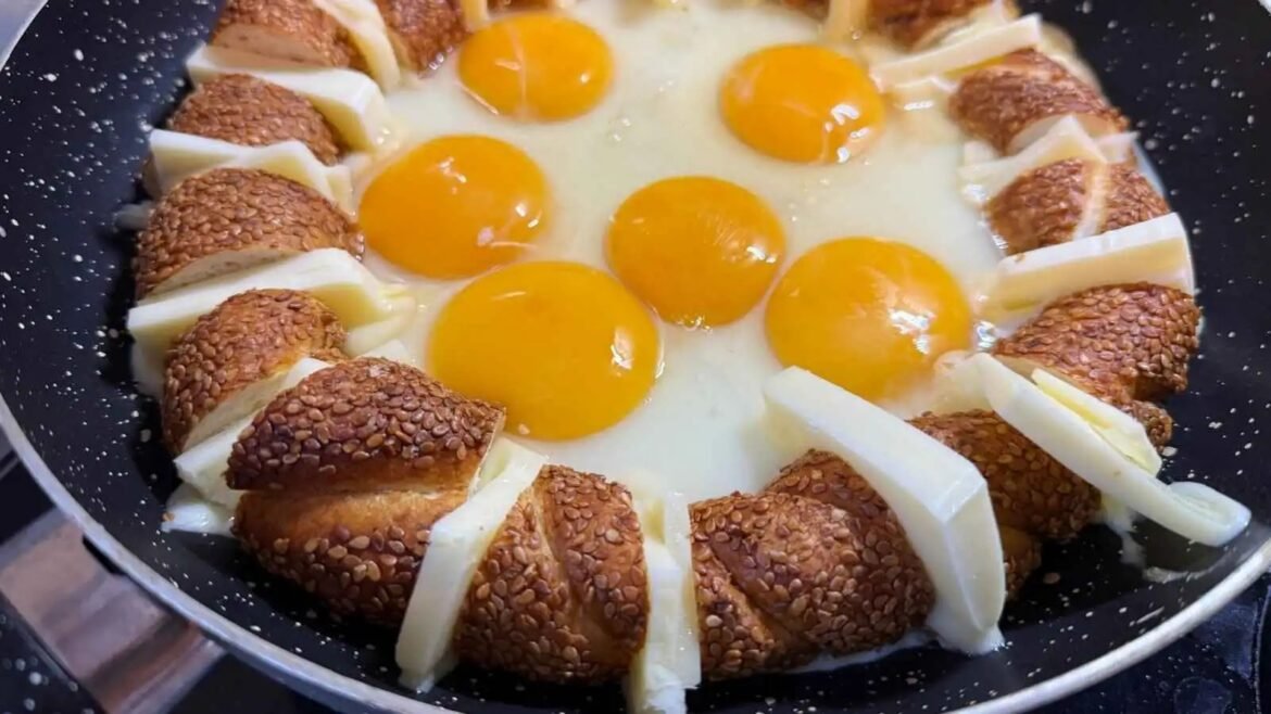 Eggs frying in a skillet arranged in a ring with sesame-crusted bread slices and cheese wedges around the edge.