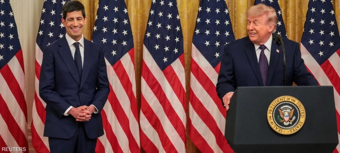 Two men in dark suits stand on a stage with multiple U.S. flags behind them; one stands at a podium with the presidential seal, smiling.
