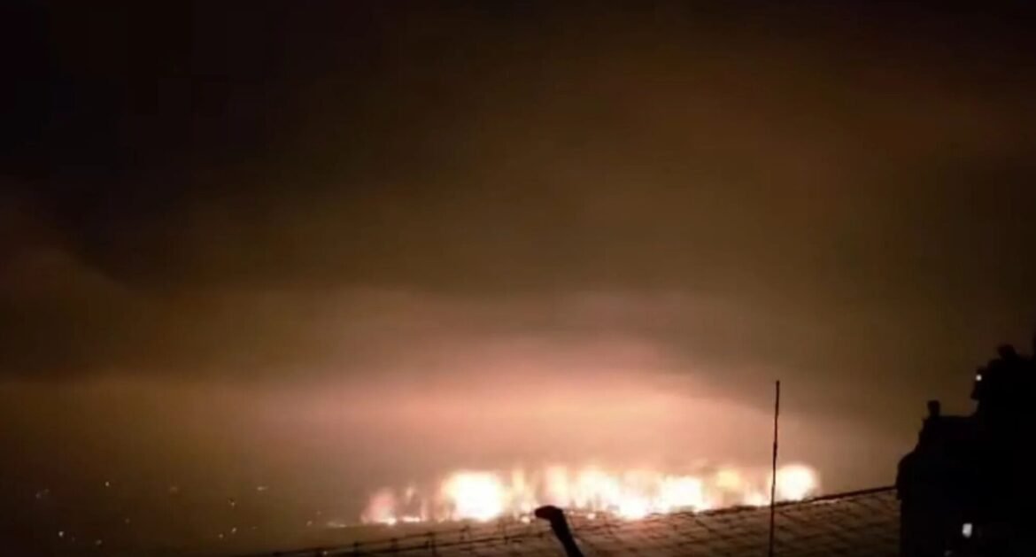 Night scene of a long line of bright flames burning along a shoreline, with smoke in the dark sky and silhouettes of a fence and buildings in the foreground.