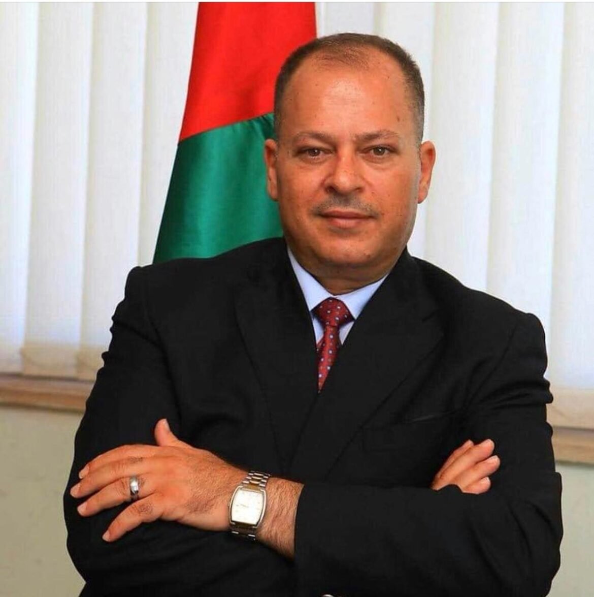 Man in a dark suit with red tie, arms crossed, in front of a red-and-green flag backdrop.
