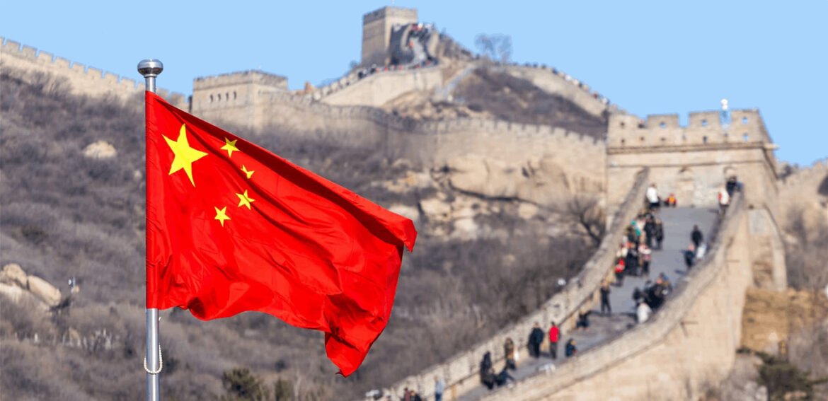 Red Chinese flag with yellow stars waving in front of the Great Wall of China and a blue sky backdrop.
