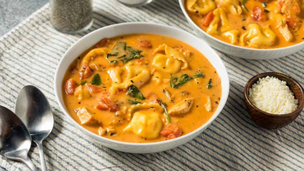 Creamy tomato-tortellini soup with spinach in a white bowl on a striped cloth; parmesan on the side.
