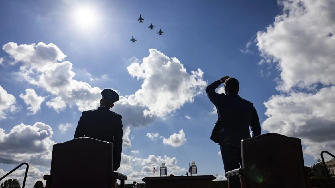 Two men, one in a military uniform, watch a formation of fighter jets fly over during a sunny ceremony.