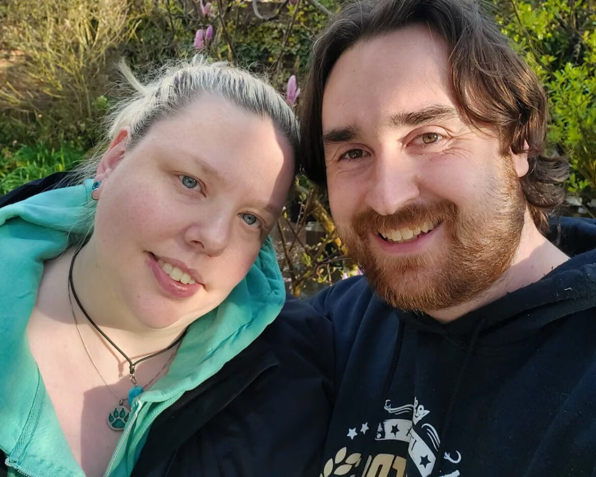 Smiling couple taking a selfie outdoors; woman in turquoise hoodie with a paw pendant, man with beard wearing a dark hoodie, greenery behind them.
