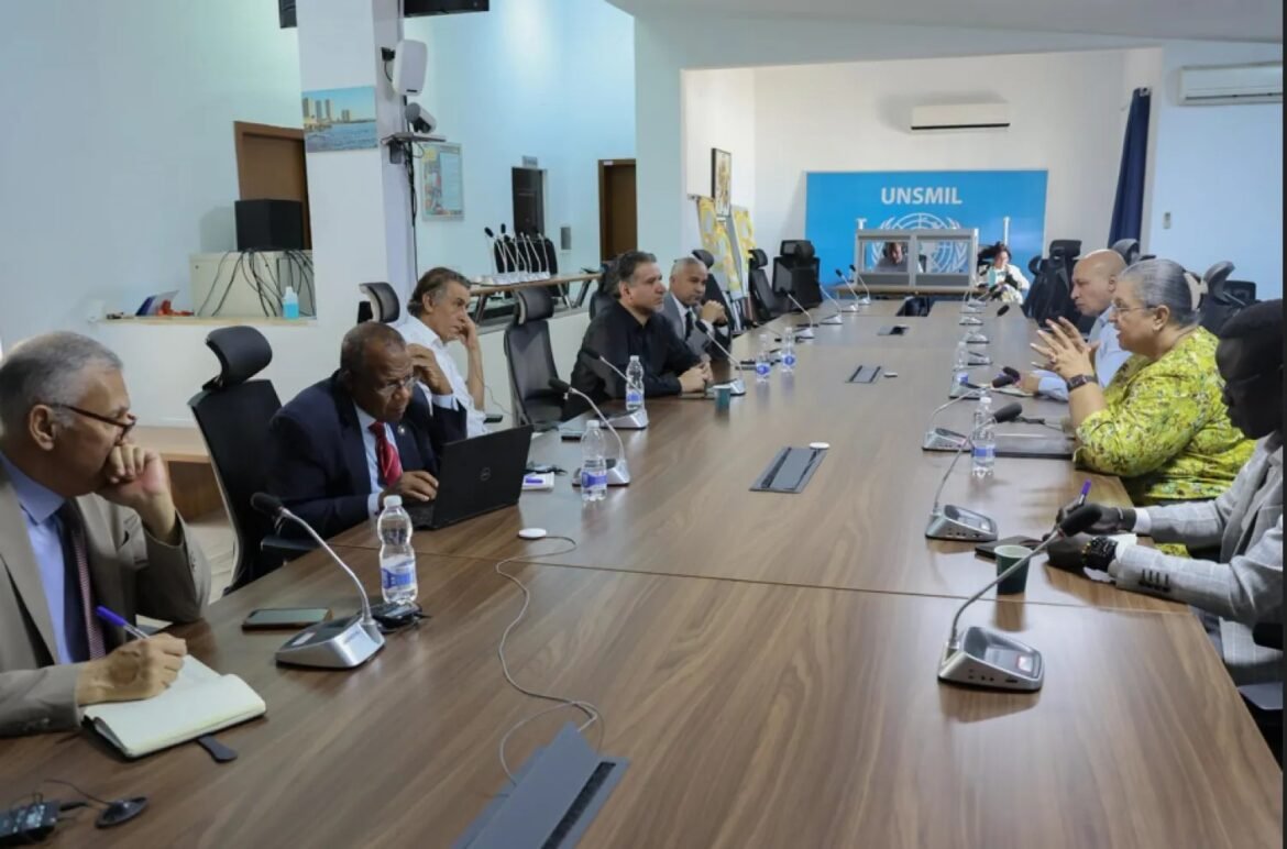 Multinational officials sit around a long wooden conference table in a formal meeting, microphones and laptops in use, with a UN UNSMIL banner in the background.