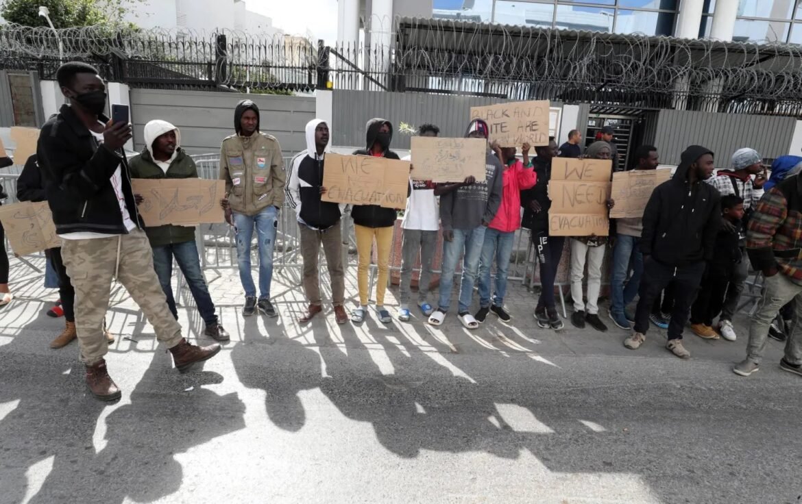 Group of protesters standing behind metal barriers, holding cardboard signs demanding evacuation outside a fenced building.