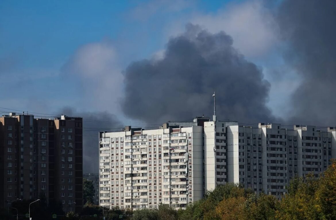 Row of gray apartment blocks with a massive dark smoke plume rising behind them in the blue sky.