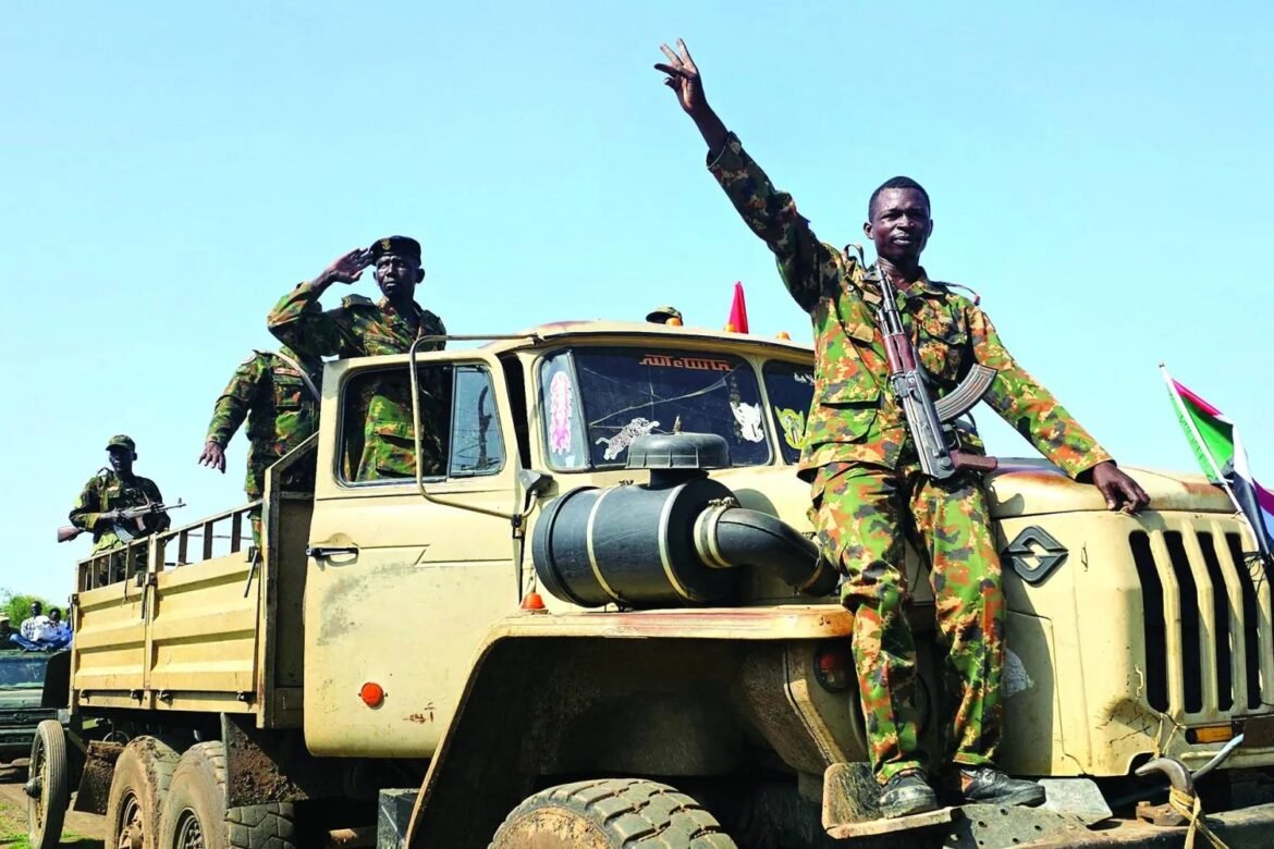 Camouflaged soldiers on a beige military truck; one officer salutes while another waves against a clear blue sky, rifle slung over his chest.