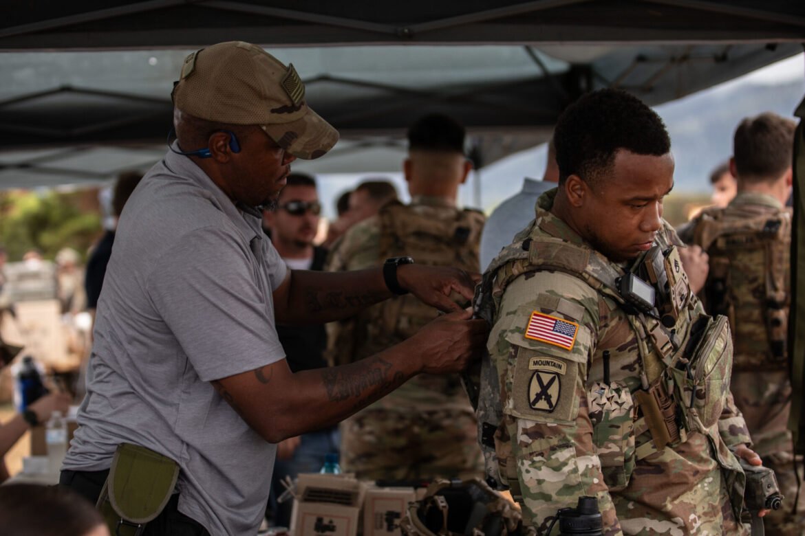 A man in a gray polo helps a soldier in camouflage gear adjust his vest under a canopy at a military event, with other soldiers in the background.