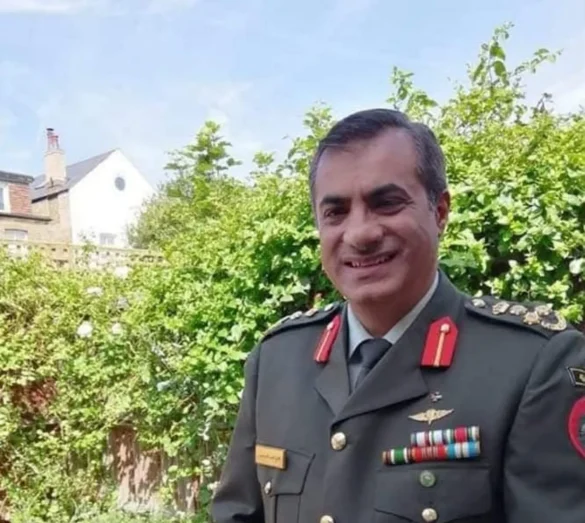Smiling man in a dark green military uniform with medals, standing outdoors among greenery.