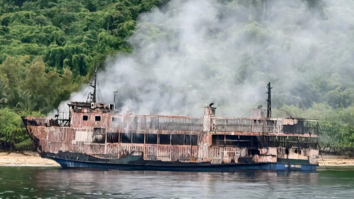Rusty, burnt-out ship along a river with smoke rising from the deck and dense green trees in the background.