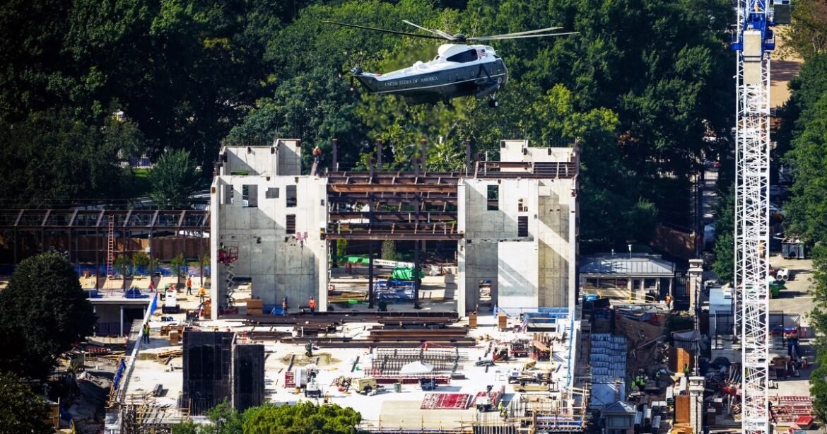 Helicopter hovers over an active construction site with a partially built concrete structure and workers on site.
