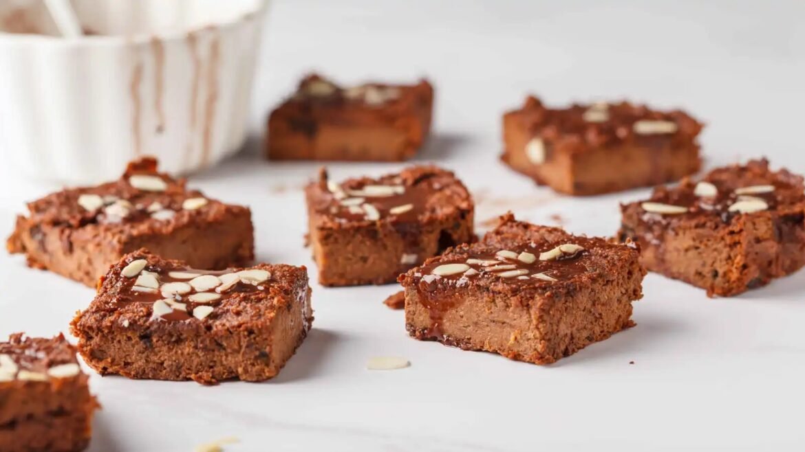 Squares of chocolate brownie bars topped with almond slices on a white surface.