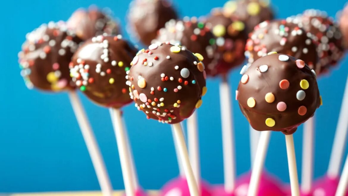 Chocolate-coated cake pops with colorful sprinkles on white sticks against a blue background.