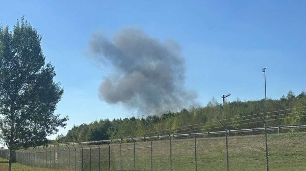 Dark gray smoke plume rising above a grassy hill behind a fence and railroad, against a clear blue sky.