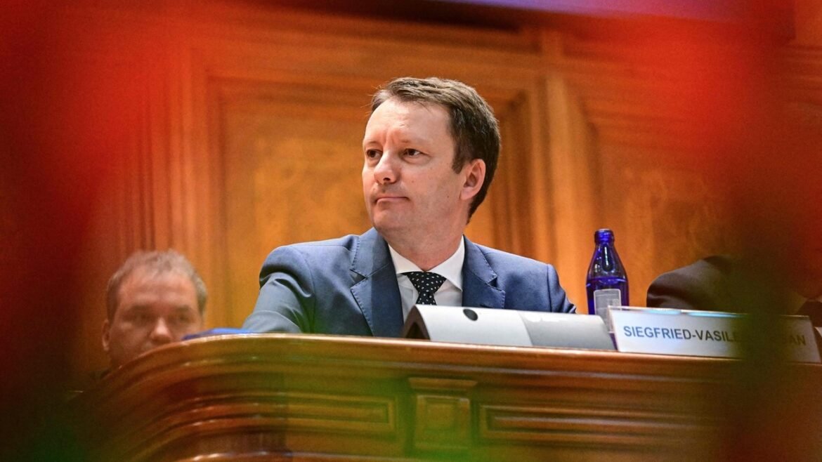 Man in a blue suit sits at a panel desk in a wood-paneled room, with a nameplate reading Siegfried-Vasilian and a blue water bottle nearby.