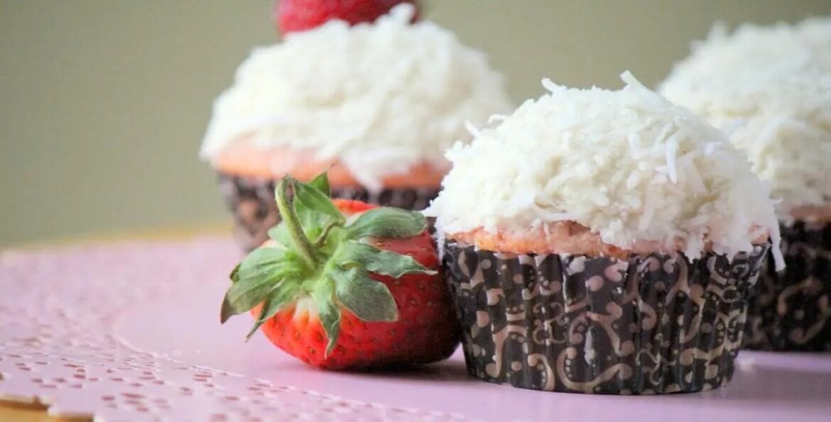 Close-up of coconut-topped cupcakes in patterned wrappers with a strawberry beside them on a pink surface.