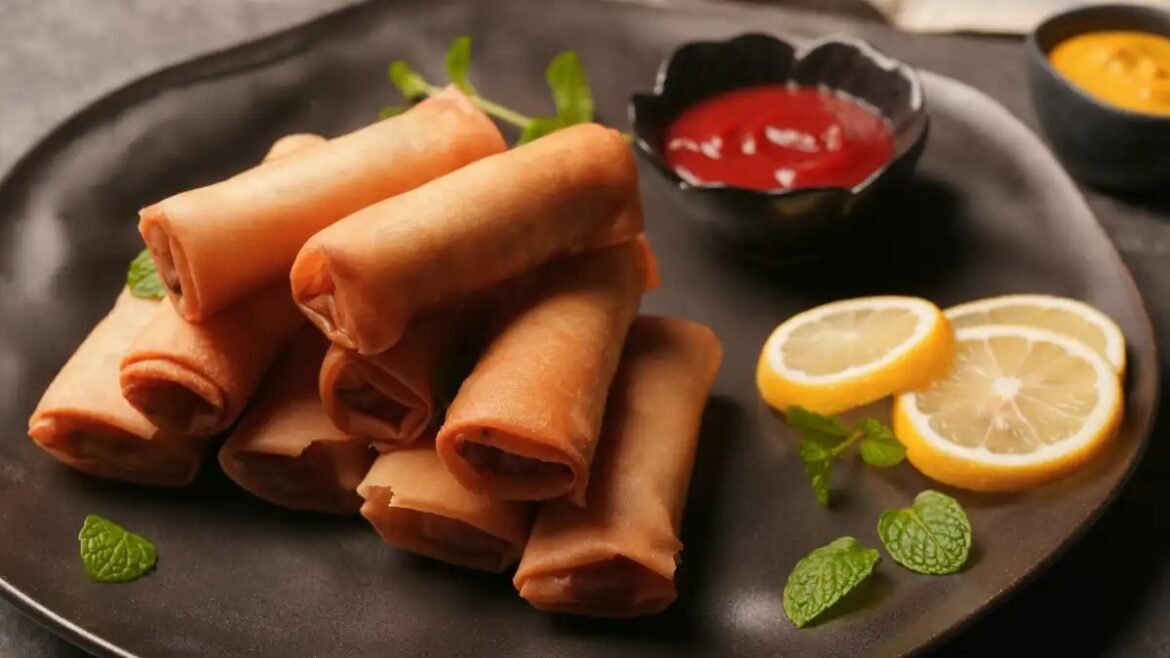Plate of fried spring rolls with lemon wedges, mint leaves, and dipping sauces on a dark platter