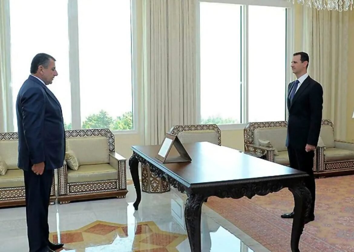 Two men in dark suits stand opposite each other in a formal office, with a large ornate desk between them and beige patterned seating nearby.