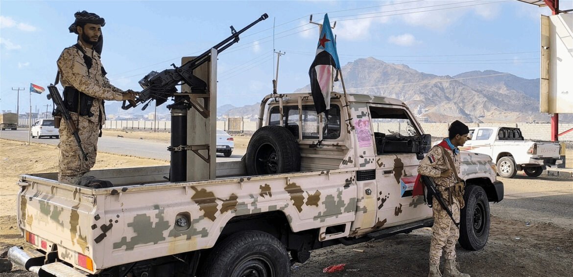 Two soldiers in desert camouflage stand by a camouflaged pickup truck with a mounted heavy machine gun in a dry, mountainous area.