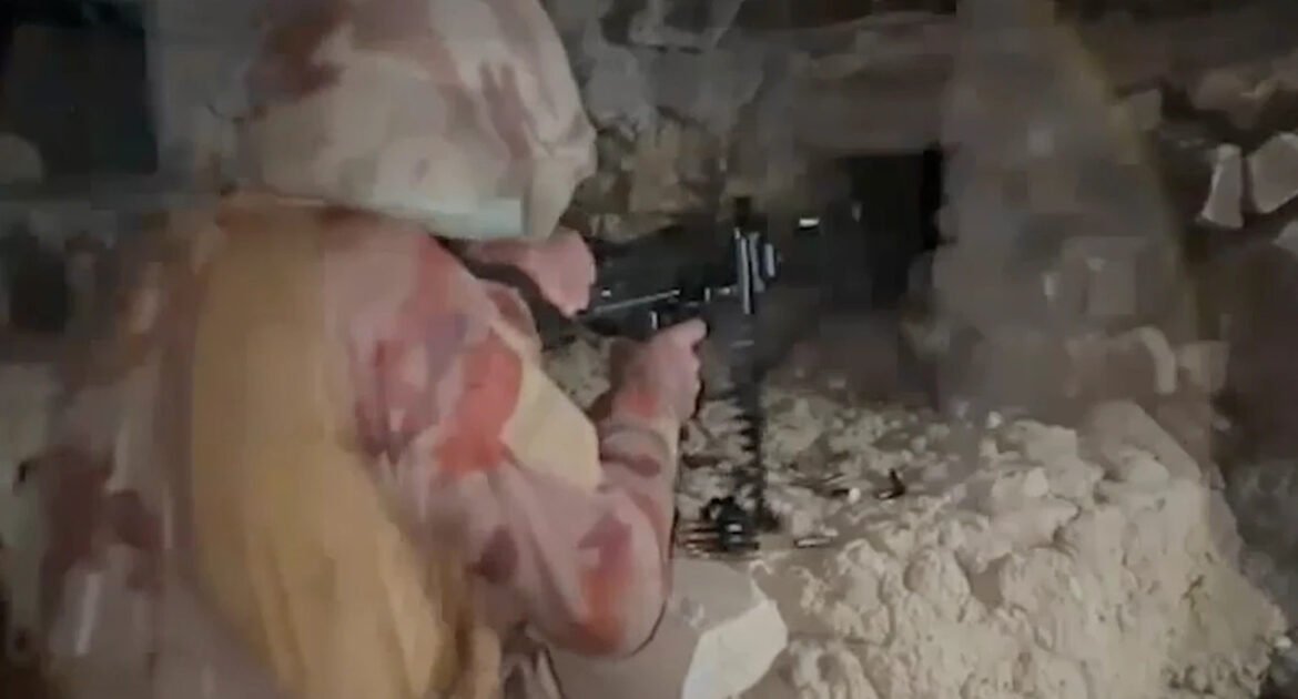Person in camouflage aiming a rifle in a rocky cave or rubble-filled tunnel at close range.
