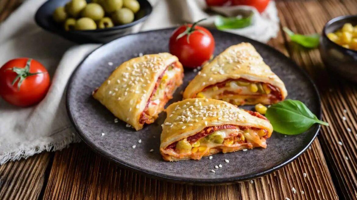 Three sesame-sprinkled pastry triangles filled with vegetables on a dark plate with tomatoes and olives nearby
