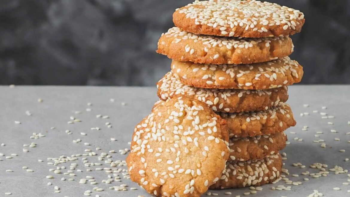 Stack of round sesame-seed cookies stacked on a gray surface with sesame seeds scattered around.