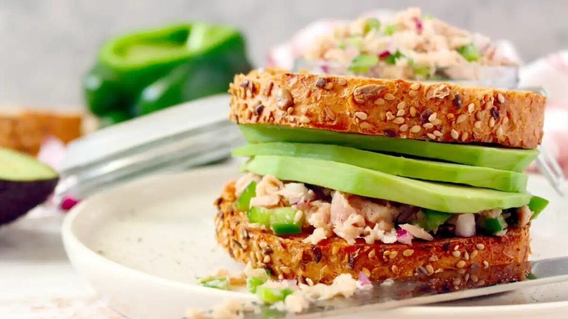 Multigrain sandwich with avocado slices and tuna salad on sesame crust, plated on a white dish.
