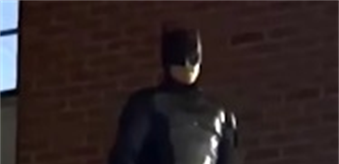 Person dressed as Batman standing against a brick wall at night, in a dark cape and cowl.