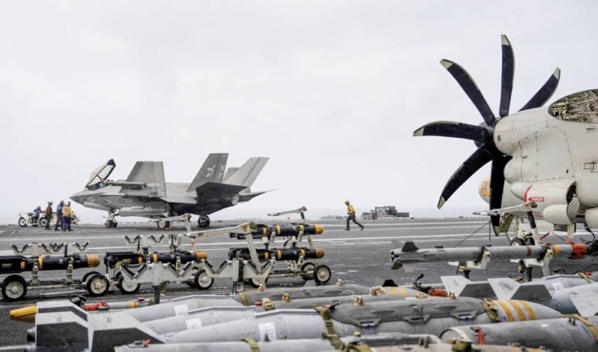 Military aircraft carrier flight deck with a parked fighter jet on the left and a large propeller aircraft on the right, surrounded by ordnance and maintenance equipment; crew in yellow vests present in the background.