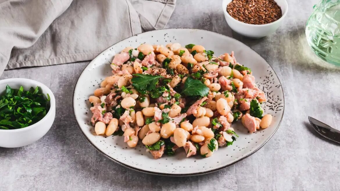 Plate of white beans with diced ham, chopped parsley, and greens in a light vinaigrette on a speckled ceramic plate necking toward a rustic table.