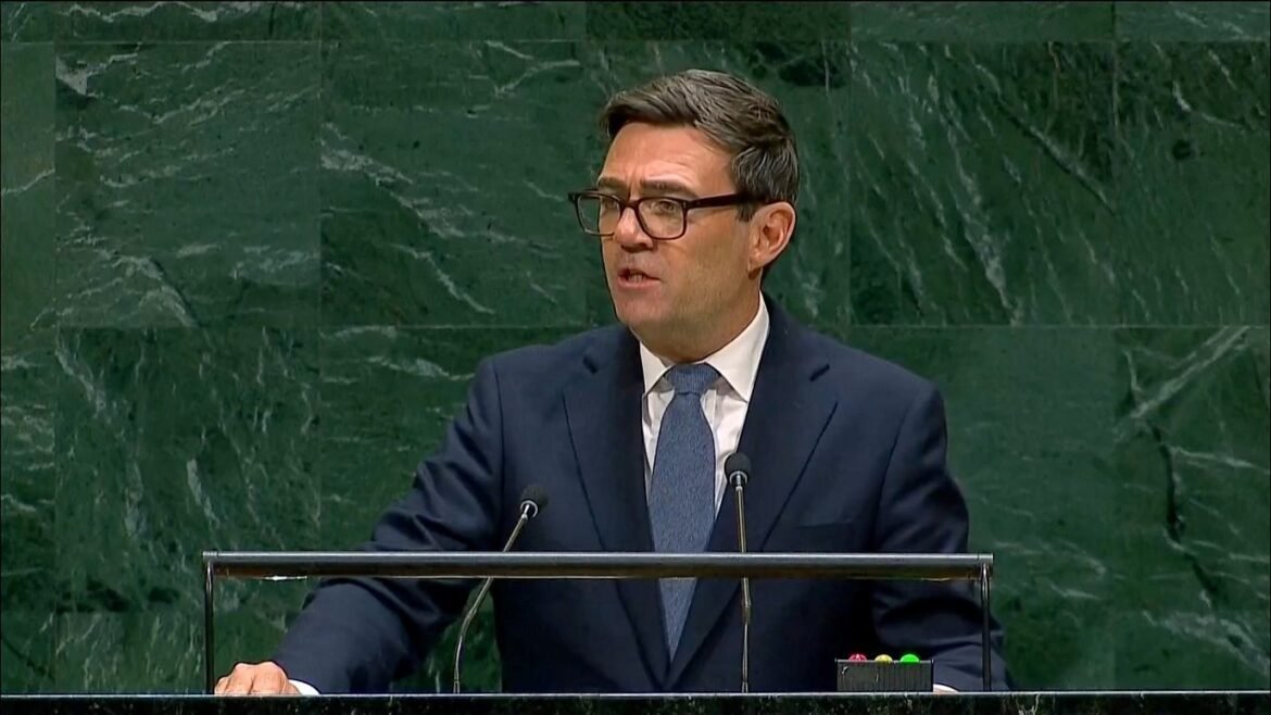 Man in a dark suit and blue tie speaks at a podium in a formal assembly chamber with a green marble backdrop and microphones in front of him.