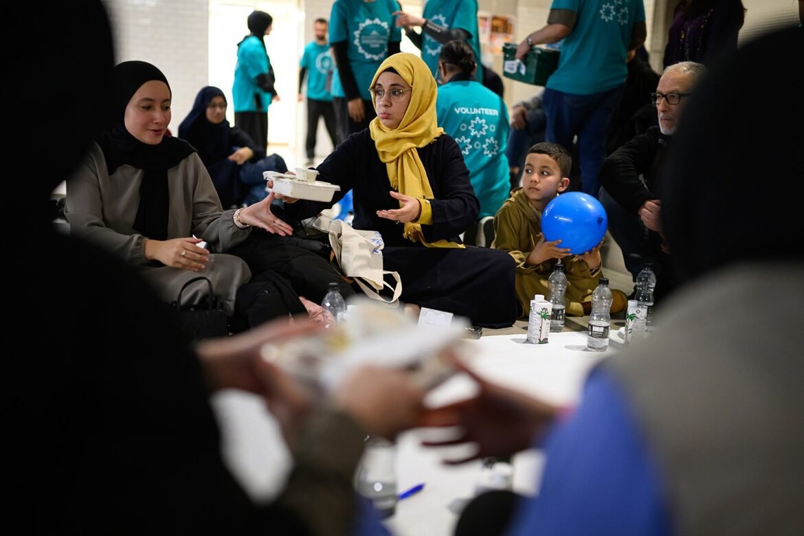 Woman in a yellow hijab passes a takeout container to a seated woman at a volunteer food event, while a boy with a blue balloon watches nearby.