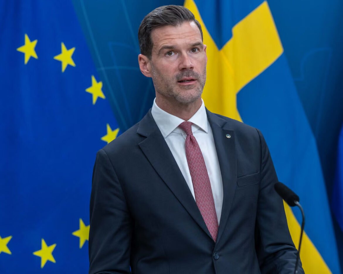 Man in a dark suit and pink tie speaks at a podium during a press conference, EU and blue-yellow flags behind him.