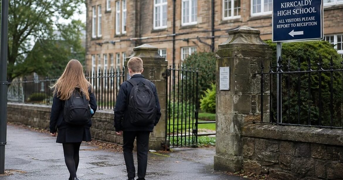 Two students with backpacks walk toward the gated entrance of Kirkcaldy High School on a wet pavement.