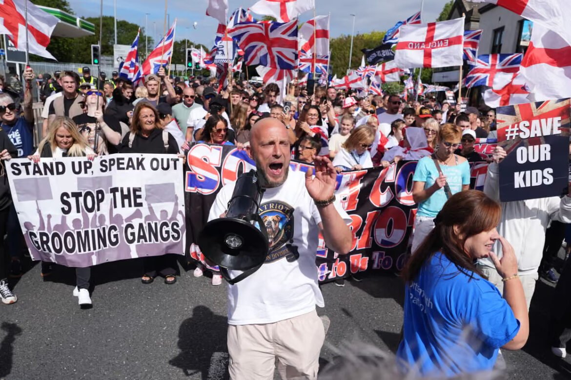 Crowd of protesters with UK flags; man with a megaphone addresses them, banners call to stop grooming gangs and save our kids