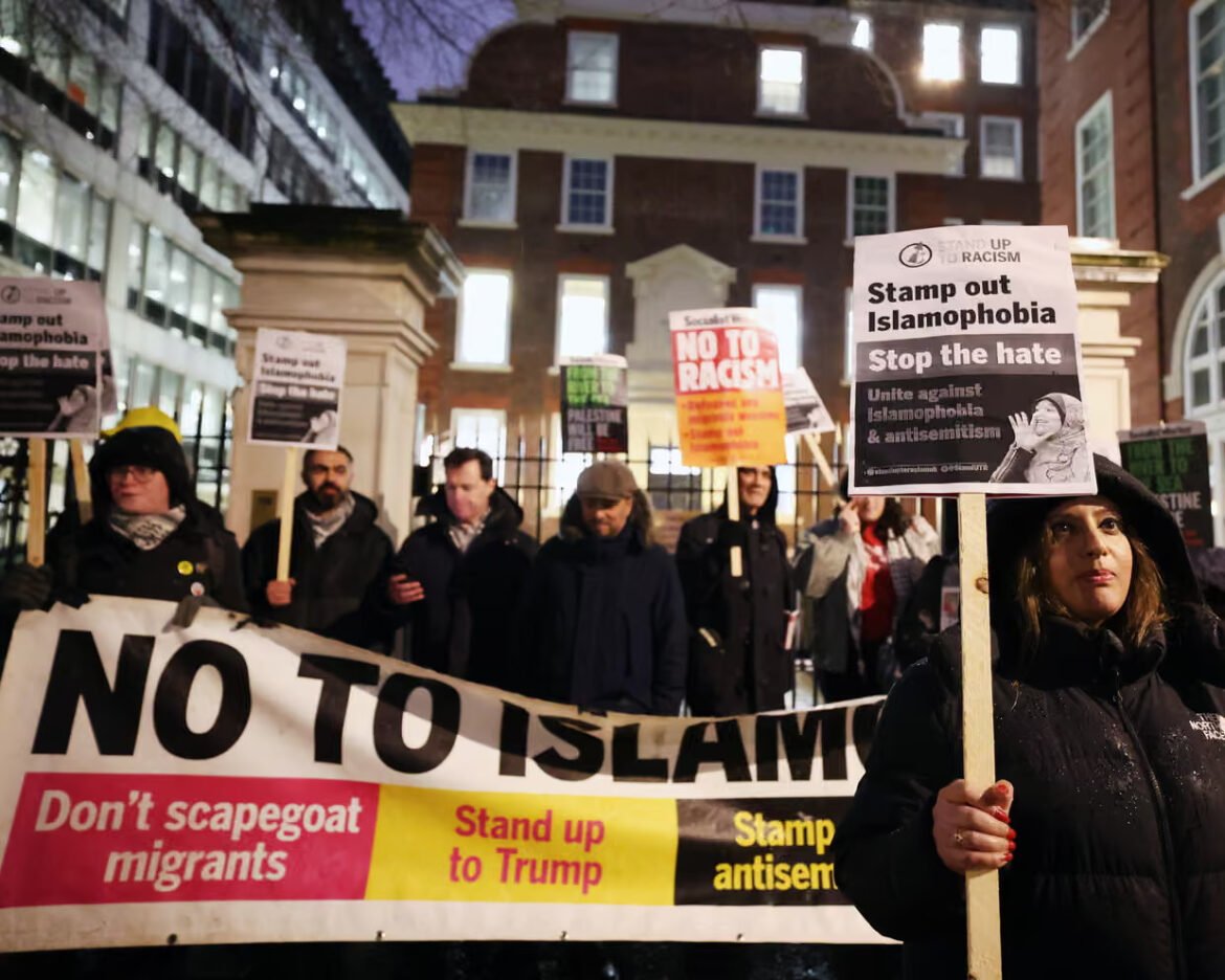 Group of protesters at night holding a large banner that reads 'NO TO ISLAM' with smaller signs including 'Stamp out Islamophobia' and 'Don't scapegoat migrants' in an urban setting.