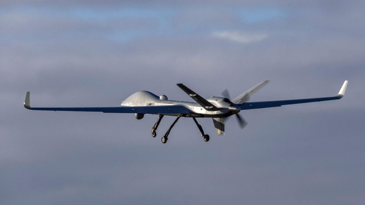 Unmanned aerial vehicle with long blue wings and a bulbous fuselage, landing gear down, in flight against a cloudy sky.
