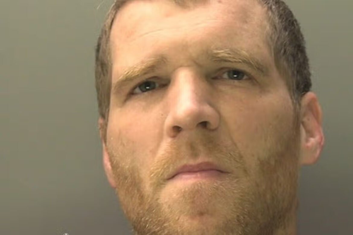 Close-up of a man with short light brown hair and light stubble, facing the camera with a neutral expression, against a plain backdrop (mugshot style).