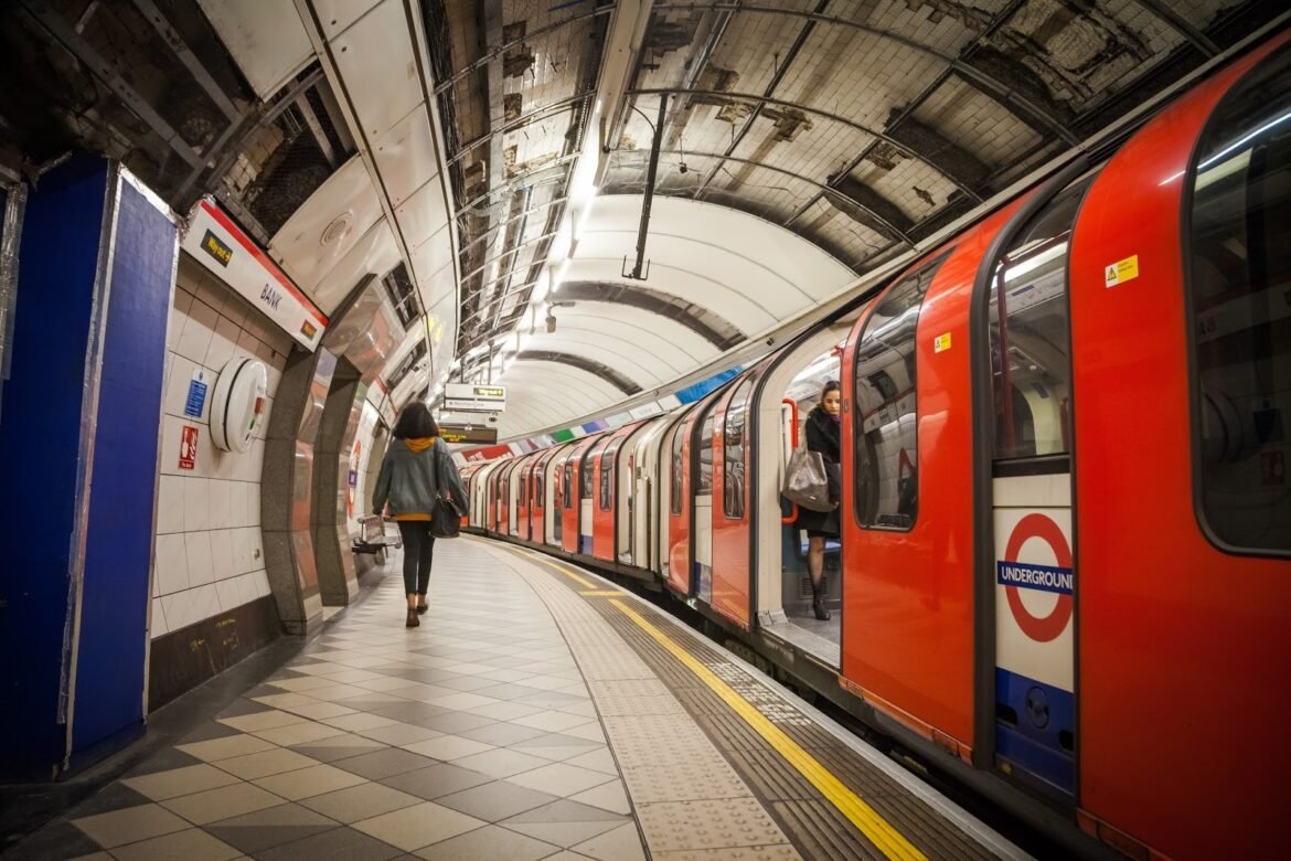 London Underground platform with a bright red Tube train at the curb; a passenger boards while another person walks along the platform with a bag.